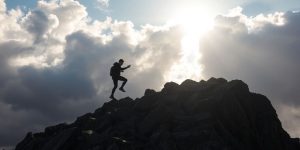 Person climbing mountain towards sunny sky.